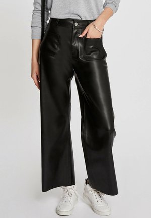 Person iført sorte vidde bukser i kunstlæder, grå sweater, hvide sneakers, med den ene hånd i lommen og sølvarmbånd på håndleddet.