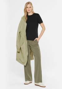 Schwarzes Kurzarmhemd kombiniert mit olivgrünen weiten Hosen und einer hellgrünen Bomberjacke. Weiße Sneakers runden das Outfit ab.