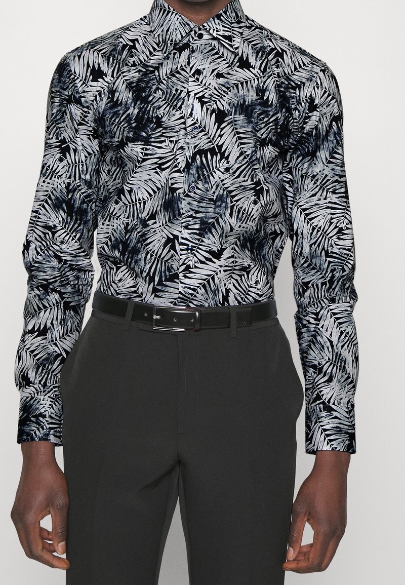 Homme portant une chemise à boutons à motif de feuilles noir et blanc rentrée dans un pantalon foncé avec une ceinture noire.