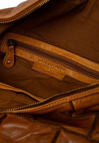 Interno di una borsa in cuoio marrone con fodera in tessuto, una tasca con zip e un'etichetta in cuoio incisa con "RE:DESIGNED EST. 2003."