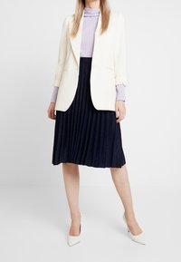 Kvinna i vit kavaj över lavendeltröja, marinblå veckad kjol och vita högklackade skor som står mot vit bakgrund.