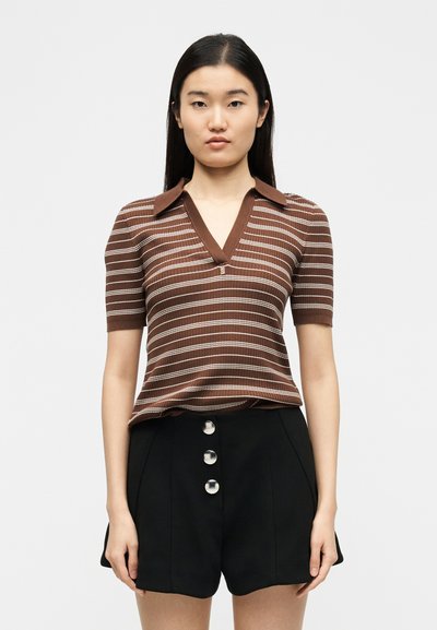 Jeune femme aux longs cheveux foncés portant un haut à manches courtes en tricot rayé marron et un short noir taille haute avec trois boutons argentés.