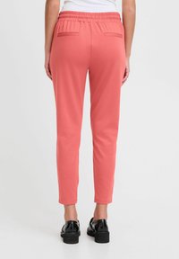 Korallfärgade byxor med elastisk midja, slim fit och två bakfickor. Materialet verkar mjukt och lätt, lämpligt för avslappnad användning.