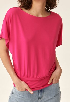 Mujer con una blusa de manga corta de color rosa brillante con cintura ajustada y jeans azul claro, con las manos en los bolsillos, frente a un fondo neutro.