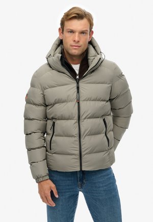 Chaqueta acolchada en gris claro con capucha, frontal con cremallera, bolsillos laterales y diseño de acolchado horizontal. Llevada sobre un suéter oscuro.