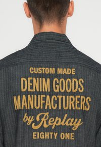 Giacca in denim scuro con motivo a spina di pesce. Presenta un ricamo dorato con la scritta "FABBRICANTI DI CAPI IN DENIM SU MISURA di Replay EIGHTY ONE."
