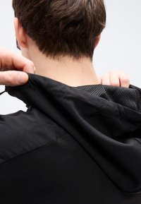 Μαύρο μπουφάν με κουκούλα, με λείο εξωτερικό και επένδυση από δίχτυ. Εστίαση στην περιοχή του λαιμού, που δείχνει την αυξομειούμενη κουκούλα και την υφή του υφάσματος.