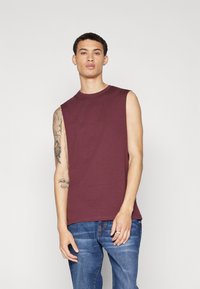 Burgunderfarget ermeløs t-skjorte i bomull med rund hals. Modellen har den på sammen med blå denimjeans, noe som viser en avslappet passform.