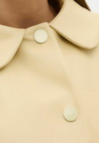 Primo piano di un cappotto beige con un ampio colletto e due grandi bottoni tondi, indossato da una persona con collo e capelli visibili.