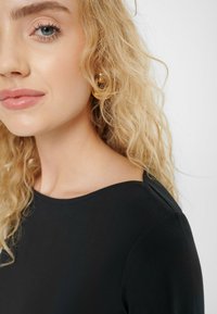 Czarna bluzka z okrągłym dekoltem, gładką fakturą i detalami przeszyć, noszona przez osobę o falujących, blond włosach.