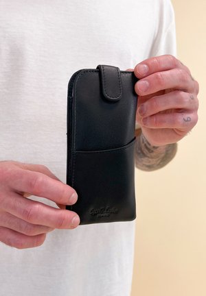 Black leather phone case with a smooth texture, rounded edges, flap closure, and a front pocket. Features embossed logo at the bottom.