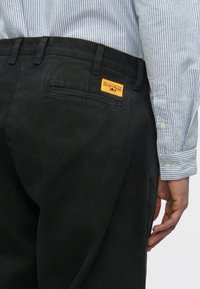 Zwarte, gestructureerde broek met een achterzak; voorzien van een opvallend oranje label met de tekst "Service Works." Het overhemd heeft lichtblauwe krijtstrepen.