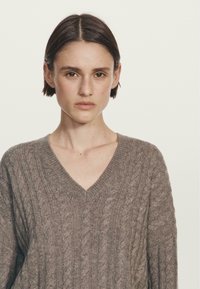 Maglione a V, dalla vestibilità ampia, in un morbido knit marrone, con un motivo a trecce testurizzato su tutta la superficie. Il tessuto appare caldo e accogliente.