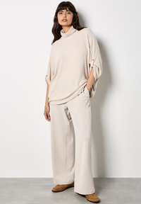 Suéter beige de cuello alto oversize con mangas enrolladas; combinado con pantalones de pierna ancha beige claro y zapatos de lona marrones. Textura suave, diseño casual.