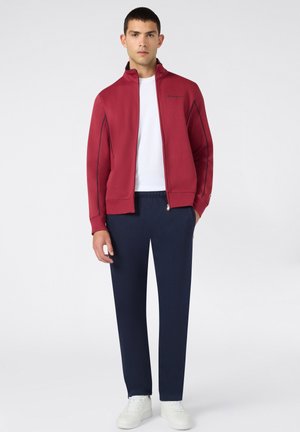Junger Mann trägt eine rote Jacke mit Reißverschluss, weißes T-Shirt, dunkelblaue Hose und weiße Turnschuhe und steht vor einem schlichten hellen Hintergrund.