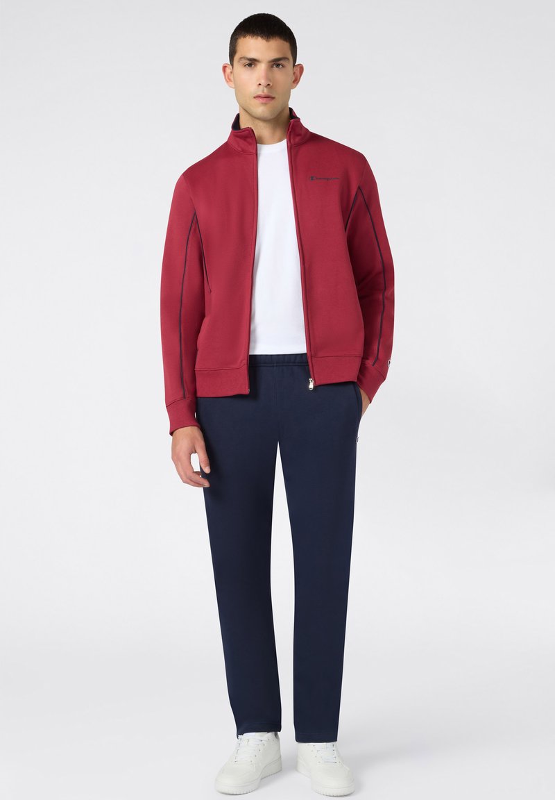 Jeune homme portant une veste zippée rouge, un t-shirt blanc, un pantalon bleu marine et des baskets blanches, debout devant un fond clair uni.