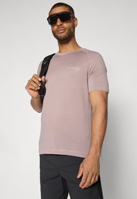 Kortermet, støvet rosa t-skjorte med teksturert stoff og en liten logo på brystet, sammen med svarte shorts og en stropp over skulderen.