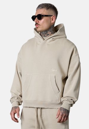 Mann mit Nacken-Tattoo und Sonnenbrille, der einen beigen übergroßen Hoodie und passende Hose trägt, steht vor einem schlichten Hintergrund.