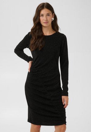 Femme aux longs cheveux bruns portant une robe noire ajustée à manches longues avec un détail froncé, debout avec une main sur la hanche devant un fond uni.