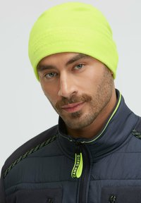 Neon yellow fleece beanie features embossed logo. Model wears a navy jacket with bright accents, textured fabric, and zippered pockets.