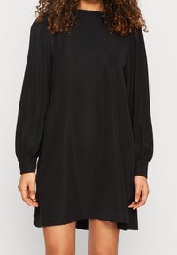 Vestido negro hecho de una tela suave, con mangas largas abullonadas, un cuello alto y un corte relajado con un dobladillo ligeramente más corto en la parte delantera.