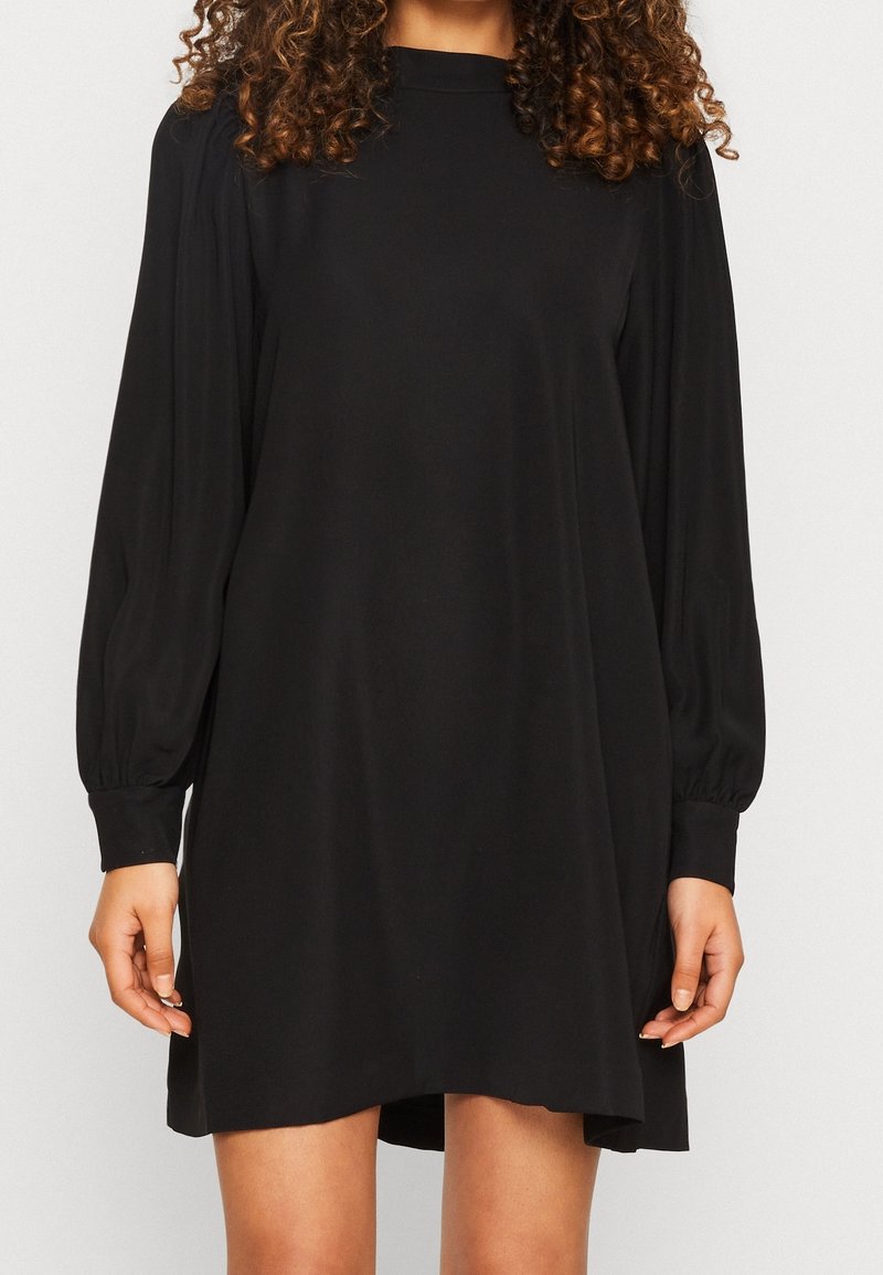 Vestido negro hecho de una tela suave, con mangas largas abullonadas, un cuello alto y un corte relajado con un dobladillo ligeramente más corto en la parte delantera.