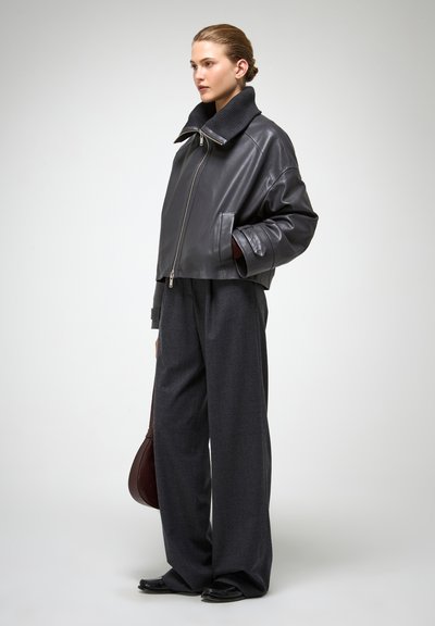 Giacca in pelle nera tagliata con colletto alto, zip argentata e due tasche laterali, abbinata a pantaloni grigi larghi e scarpe nere.