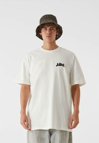 Cămașă albă din bumbac cu mâneci scurte, având un grafic negru pe piept. Asortată cu o pălărie de tip bucket texturată, de culoare verde măslinie.