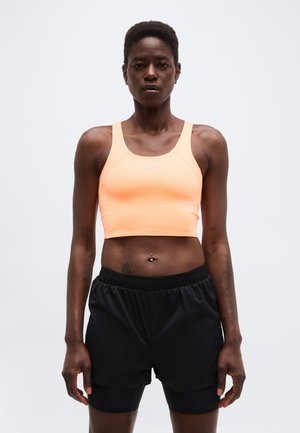 Personne athlétique portant un débardeur de sport court pêche et un short d'entraînement noir en couches, debout devant un fond blanc uni.