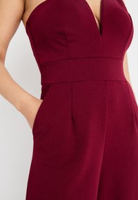 Salopette bordeaux con scollatura a V profonda, busto strutturato, vita larga e tasche laterali. Tessuto liscio con finitura opaca.