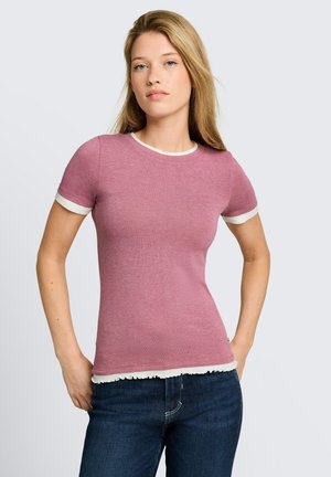 Jonge vrouw met lang blond haar, gekleed in een nauwsluitend roze shirt met korte mouwen en witte bies en donkerblauwe spijkerbroek, staand met handen in de heupen.