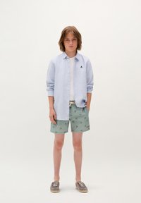 Camicia a righe azzurra chiara con bottoni, t-shirt bianca, pantaloni corti verdi con motivo floreale e espadrillas grigie con lacci.