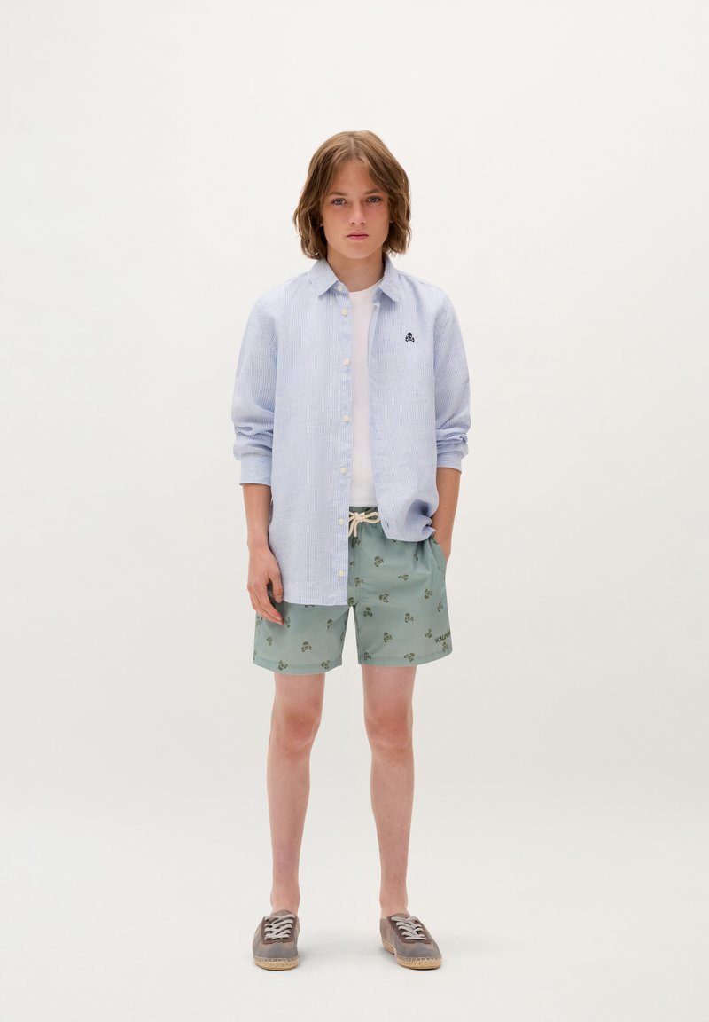 Camicia a righe azzurra chiara con bottoni, t-shirt bianca, pantaloni corti verdi con motivo floreale e espadrillas grigie con lacci.