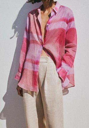 Kvinde iført en løs, langærmet skjorte med pink og røde striber og lyse beige højtaljede viddebukser, stående mod en hvid væg.