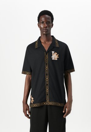 Zwart shirt met korte mouwen, voorzien van gouden borduursels langs de kraag en de knopenlijn, met een "LA" logo en bloemdetail aan de zoom.