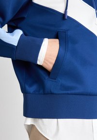 Main insérée dans la poche avant d'un sweat à capuche bleu à blocs de couleur avec détails blancs et bleu clair, par-dessus une chemise blanche et un pantalon beige.