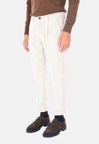 Pantaloni a gamba affusolata di colore chiaro con pieghe, dotati di una vita elastica e chiusura con bottone, abbinati a scarpe in suede marrone scuro.