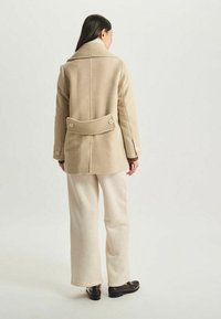 Manteau en laine beige au design ajusté, grande collerette et détail boutonné à l'arrière, associé à un pantalon large crème et des chaussures sombres.