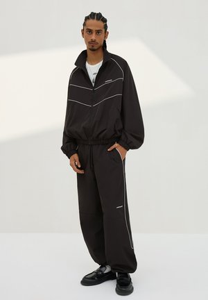 Man met gevlochten haar, gekleed in een zwart trainingspak met witte strepen, zwarte schoenen, staand met één hand in de zak tegen een effen lichte achtergrond.