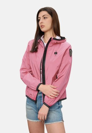 Giovane donna con lunghi capelli castani che indossa una giacca rosa con cerniera e bordi neri e pantaloncini di jeans blu, posando davanti a uno sfondo semplice.