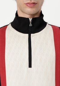 Halbzip-Pullover in Creme mit geometrischer Textur; schwarzer Kragen und rote Seitenpanele. Mit silbernem Reißverschluss und auffälliger Zuglasche.