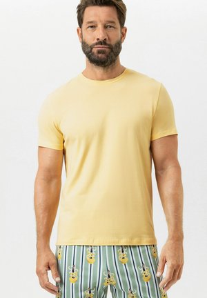 Mann mit Bart, der ein schlichtes gelbes Kurzarm-T-Shirt und gestreifte Shorts mit gelben und marineblauen Mustern trägt, steht vor einem weißen Hintergrund.