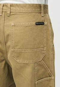 Κοντινό πλάνο στην πίσω τσέπη χακί παντελονιών cargo με ετικέτα Quiksilver και λεπτομερή λευκή ραφή σε ανθεκτικό ύφασμα.