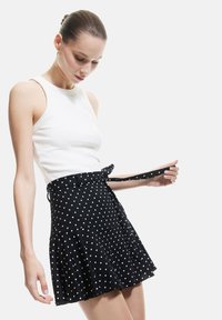 Gonna nera a pois con un design svasato, realizzata in tessuto testurizzato. Presenta una cintura da annodare e una vita aderente. Abbinata a una canotta bianca.