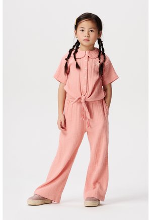 Roze katoenen tweedelige outfit met korte mouwen, kraag, trekkoord in de taille, brede broek en gestructureerde stof. Model staat met handen in de zakken.