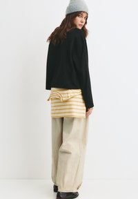 Maglietta nera a maniche lunghe cropped indossata sopra una camicia a righe gialle e bianche, abbinata a pantaloni beige a gamba larga e un berretto grigio chiaro.
