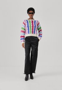 Mehrfarbiger Pullover mit strukturierten Streifen in Pink, Grün, Blau und Weiß, kombiniert mit glänzenden schwarzen Cargo-Shorts und schwarzen Stiefeletten.