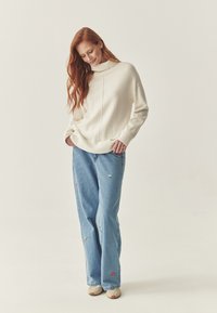 Maglione bianco lavorato a maglia con orlo a coste e colletto alto, abbinato a jeans azzurri chiari con ricamo floreale colorato. Vestibilità casual e texture visibile.