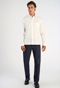 Man in een wit overhemd met knopen, donkerblauwe broek en zwartleren schoenen, staande tegen een effen lichte achtergrond.