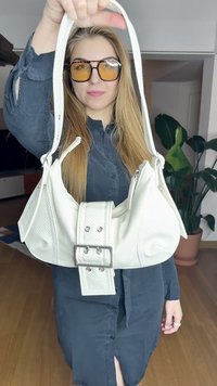 Hvid håndtaske med en tekstureret finish, der har en bred spænde rem og korte skulderhåndtag, holdt i hånden mod en neutral baggrund.